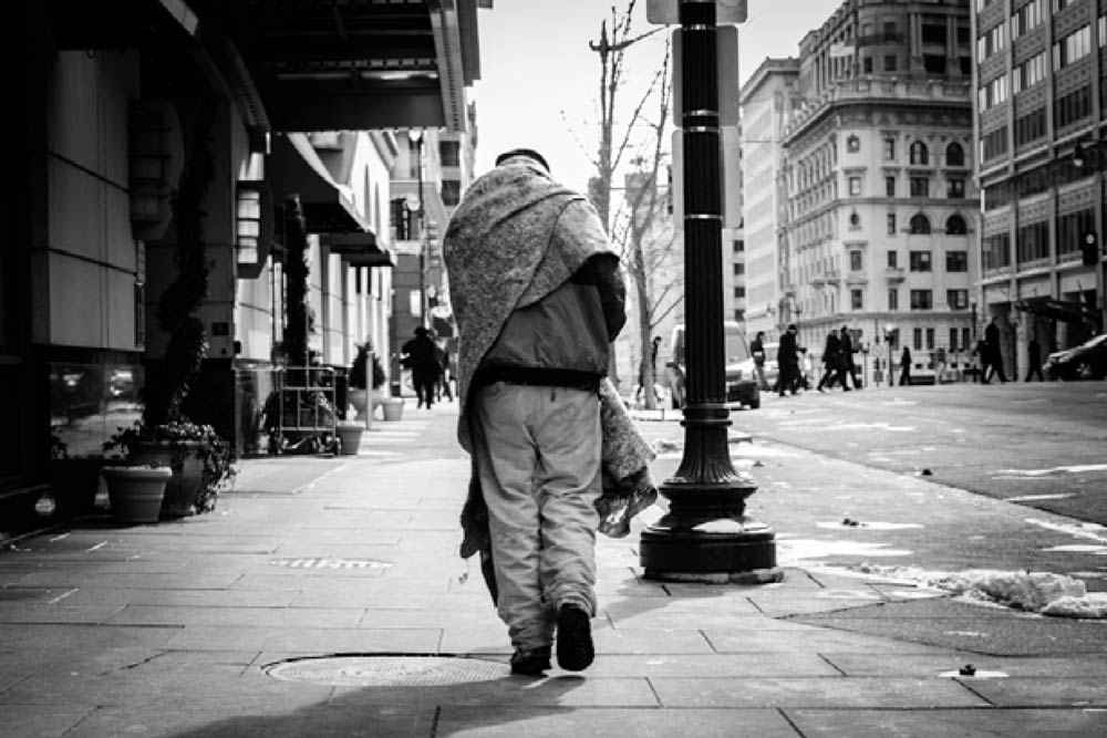 Black and white photo of man walking downtown, slumping with a heavy blanket over his shoulders. He is presumably homeless.