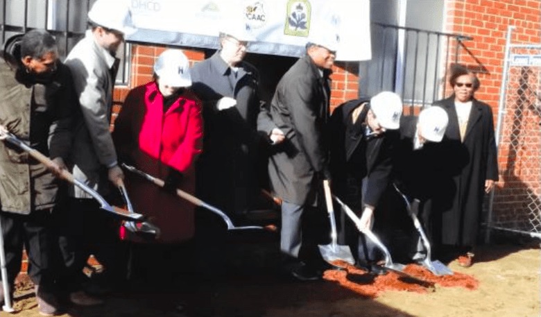 Color photo of city, state, and county officials with shovels and wearing hard hats for a ceremonial groundbreaking