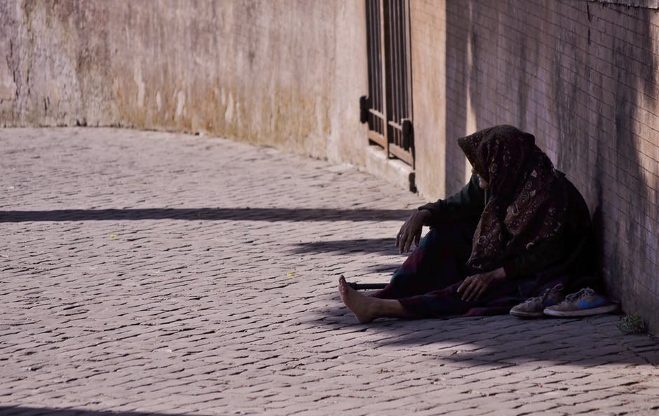 Color photo of a person, who appears to be experiencing homeless, sitting on the ground against a wall with a hood up over their head
