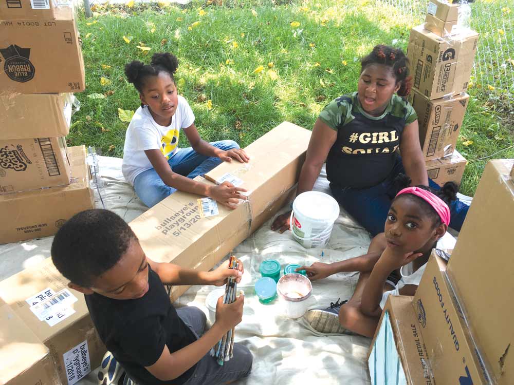 Photo of four children sitting in a circle with some cardboard building supplies on a blanket outside.