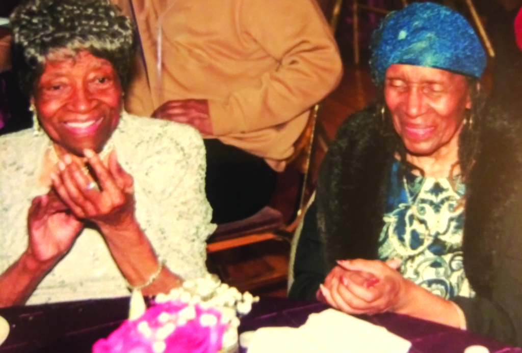 Color photo of two older women sitting at a table, on in a white coat and the other wearing a blue blouse and matching blue hat