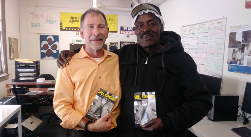 COlor photo of two men standing with their arms over each other's shoulders, holding books in front of them