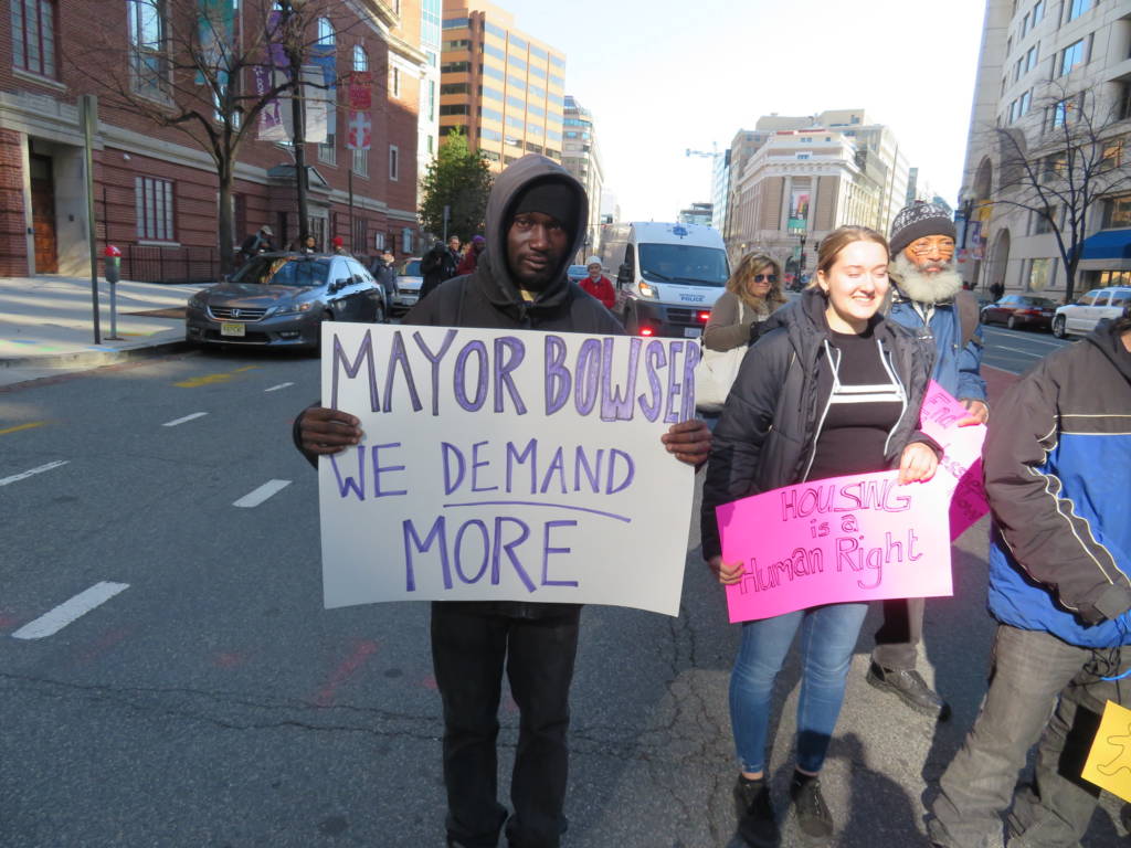 Photo of a man holding a sign that says "Mayor Bowser, we demand more." The woman next to him holds a sign that says "Housing is a human right."
