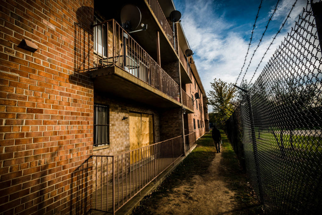 Photo of a dirt path running between a brick apartment building with plywood panels over the doors and a fence with barbed wire on the top.
