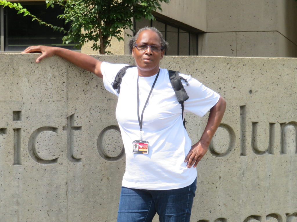 A photo of Sheila White leaning against a University of the District of Columbia sign and wearing her student ID