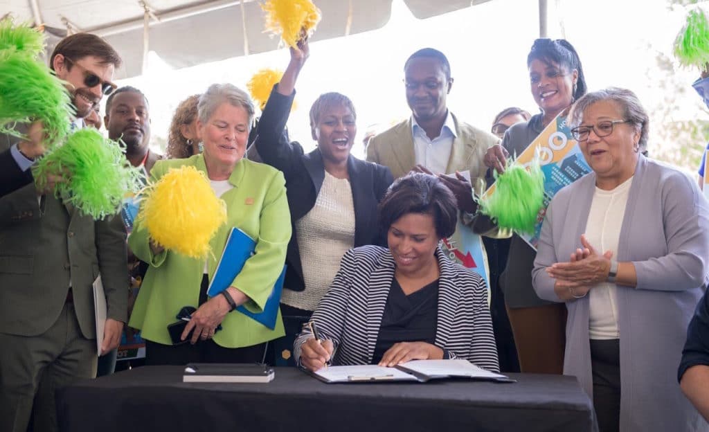 A photo showing a celebratory rally with DC Mayor Muriel Bowser signing a new executive order for housing, surrounded by many people and supporters.
