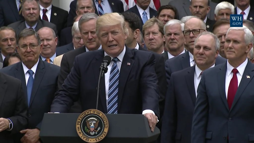 Photo of President Trump at a podium with old white men in suits.