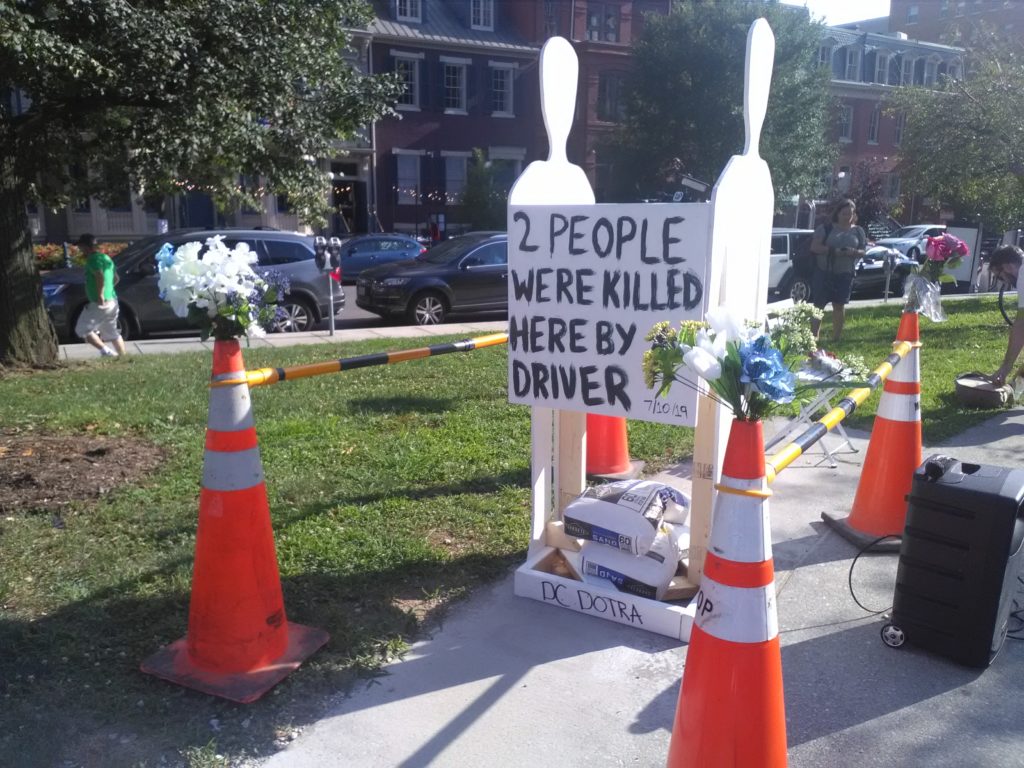 Photo of a display at the vigil: two cut-out figures of people, surrounding by traffic cones and flowers, with a sign that says "2 people were killed here by a driver"