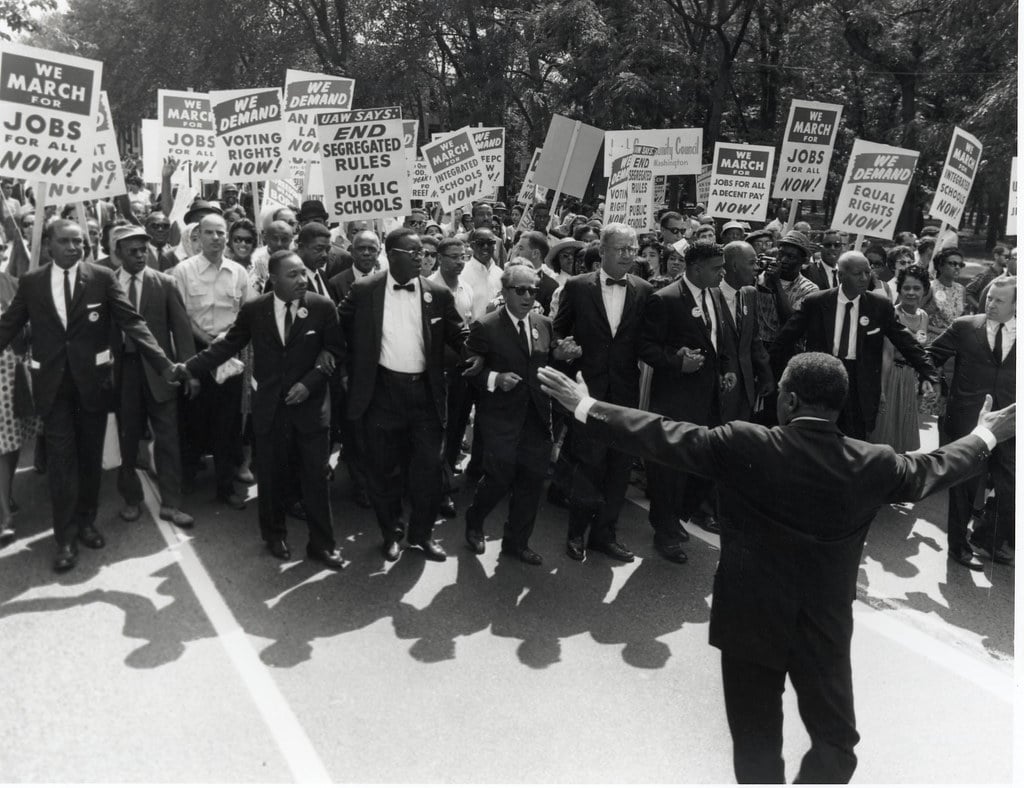 Black and white photo of MLK leading a crowd of demonstrators carrying protest signs.