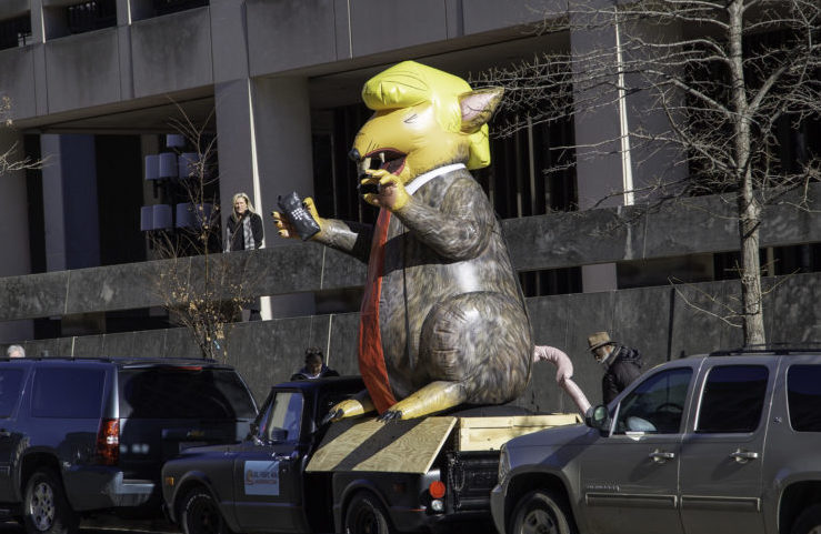 A giant blow-up rat with yellow hair and a long red tie, holding a wireless phone handset outside the federal courthouse.
