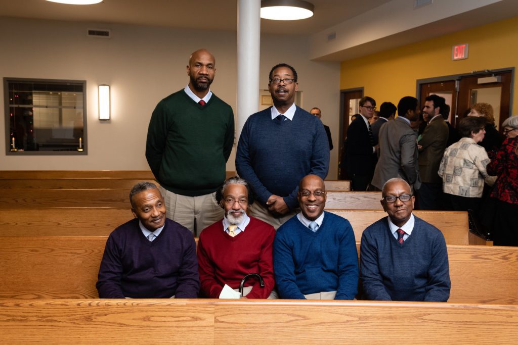 A photo of 6 black men. Four of the men sit down in a church pew with the other two men standing behind them. All wear sweaters with collared shirts and ties.