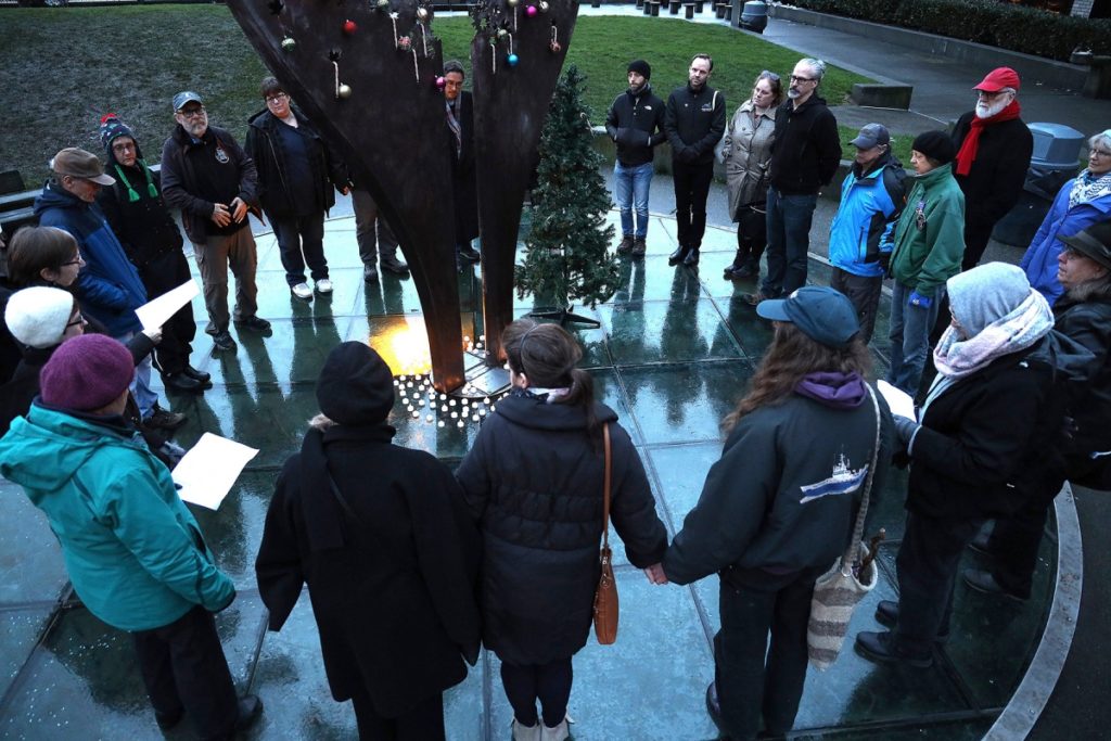 A circle of people holding hands surround a memorial that has a small Christmas tree placed next to it and ornaments hung on it