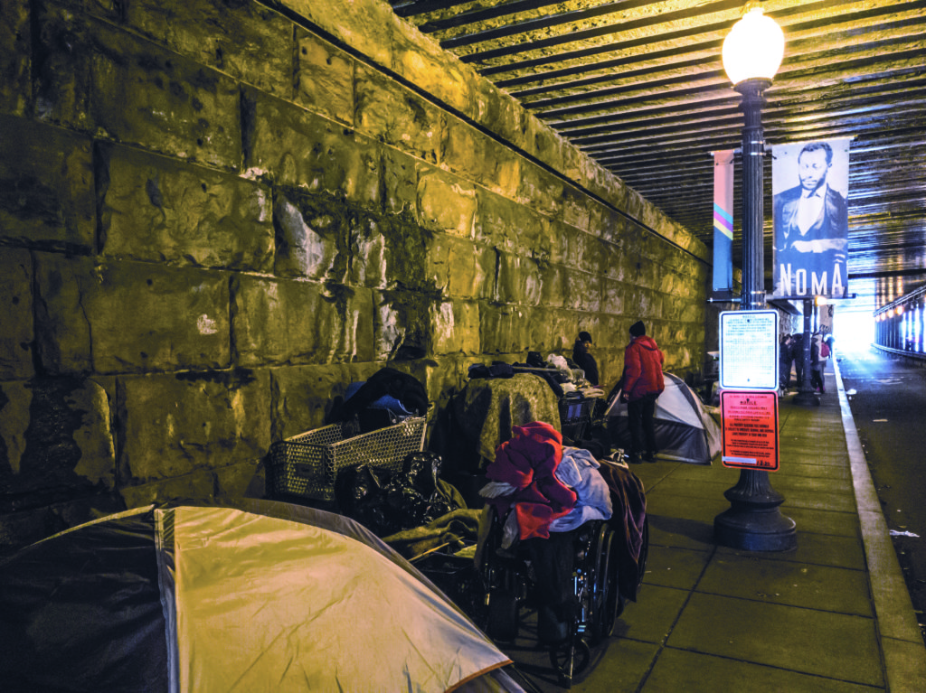 Homeless residents under a bridge in NoMa stand next to their tents and belongings before the clean up process begins. A sign advertising the clean-up process hangs on a light post nearby.