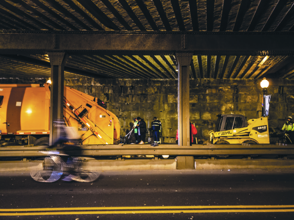 A front loader follows behind city workers and a Department of Public Works truck as they sweep the sidewalk and throw items left in the K Street underpass.
