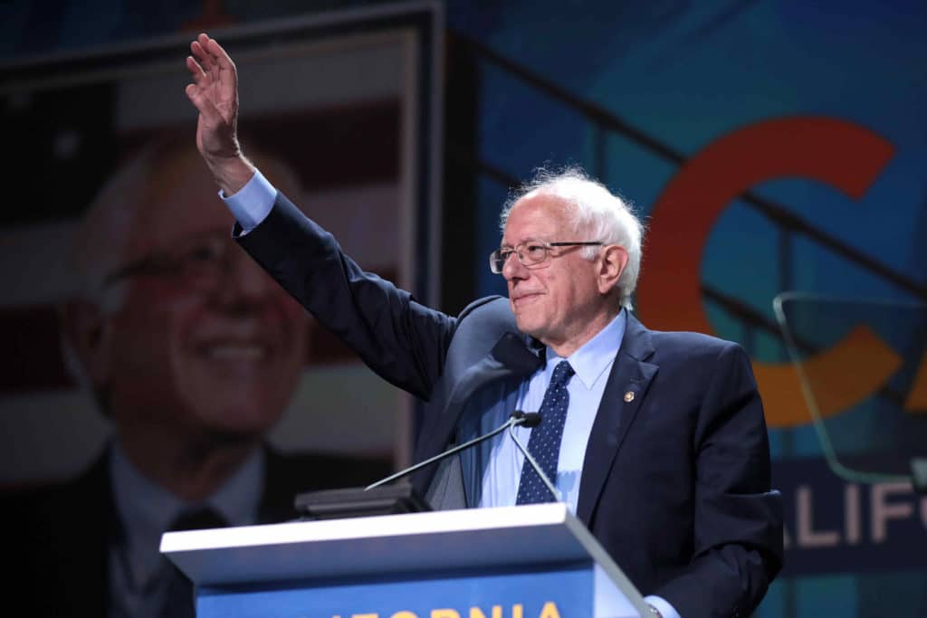 Bernie Sanders stands at a podium, wearing a suit and waving.
