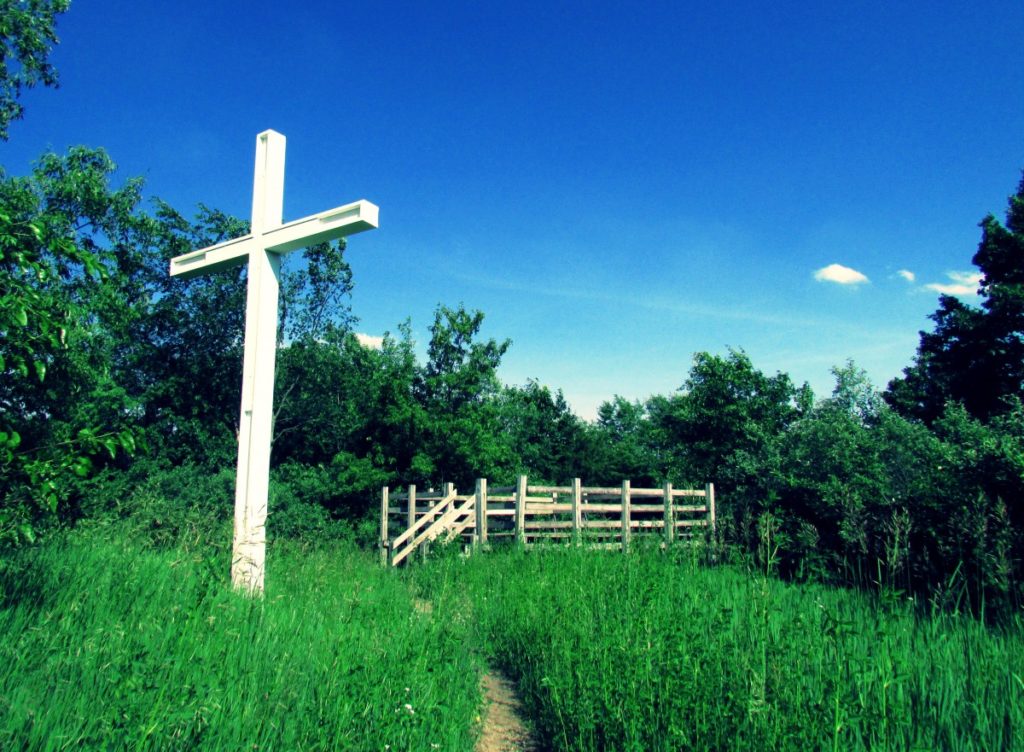 A cross stands near a white picket fence in a rural area.
