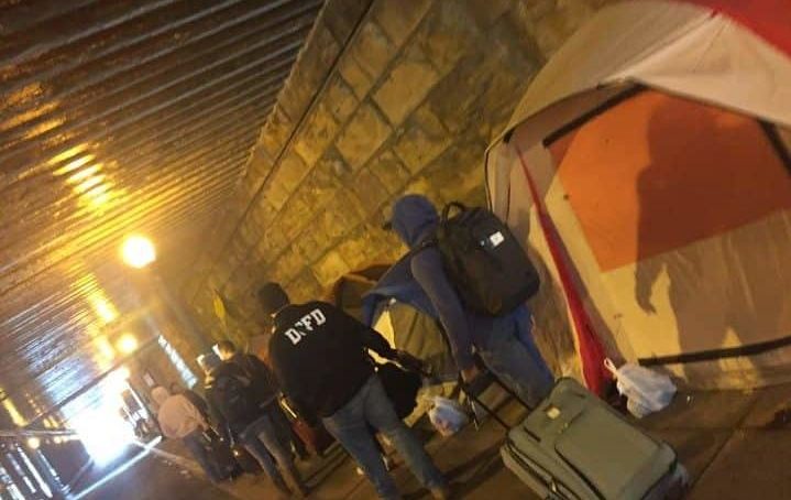 Photo of a line of people carrying supplies next to several tents in a dark tunnel.