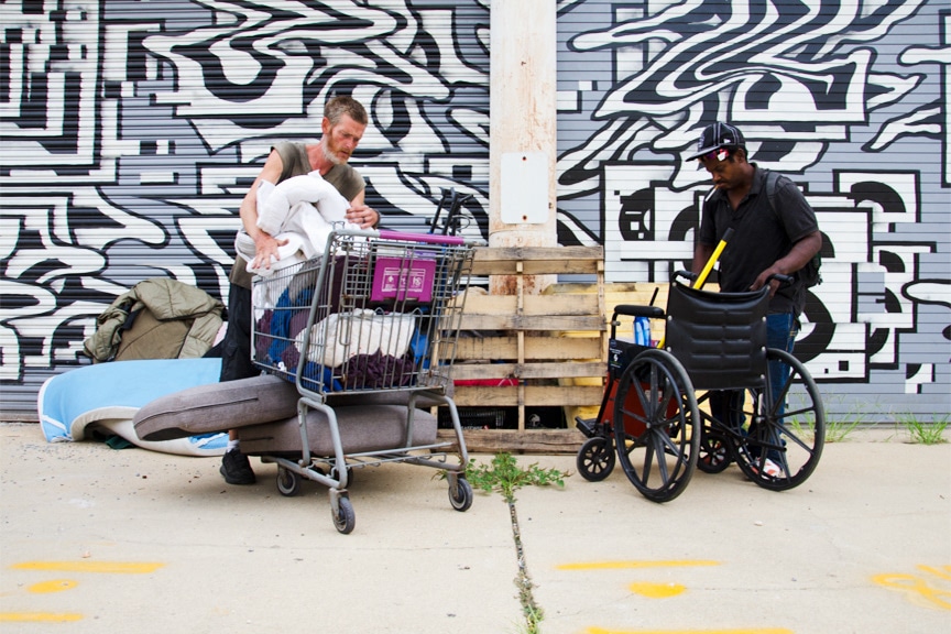 Photo of two men experiencing homeless who are moving their belongings in a shopping cart and a wheelchair.