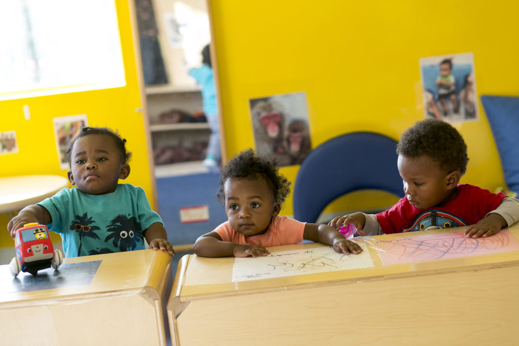 Three African-american toddlers sit at a classroom table and use crayons on a sheet of