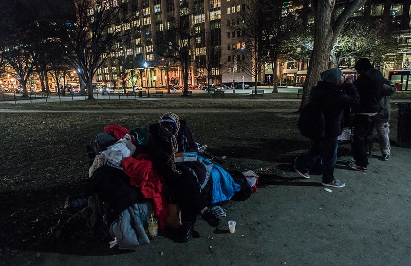 Photo of two people walking towards a man experiencing homelessness who is sitting on a park bench at night.