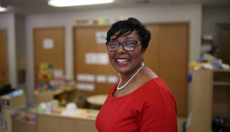 Black woman smiling at the camera wearing a red dress and pearls. Background is out of focus and depicts a classroom.