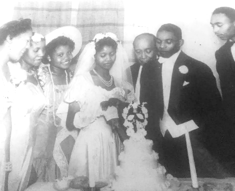 In a black and white photo, a black family is dressed up for a wedding. The bride is wearing a dress and a veil and looking at a four tier cake with a bow on top.