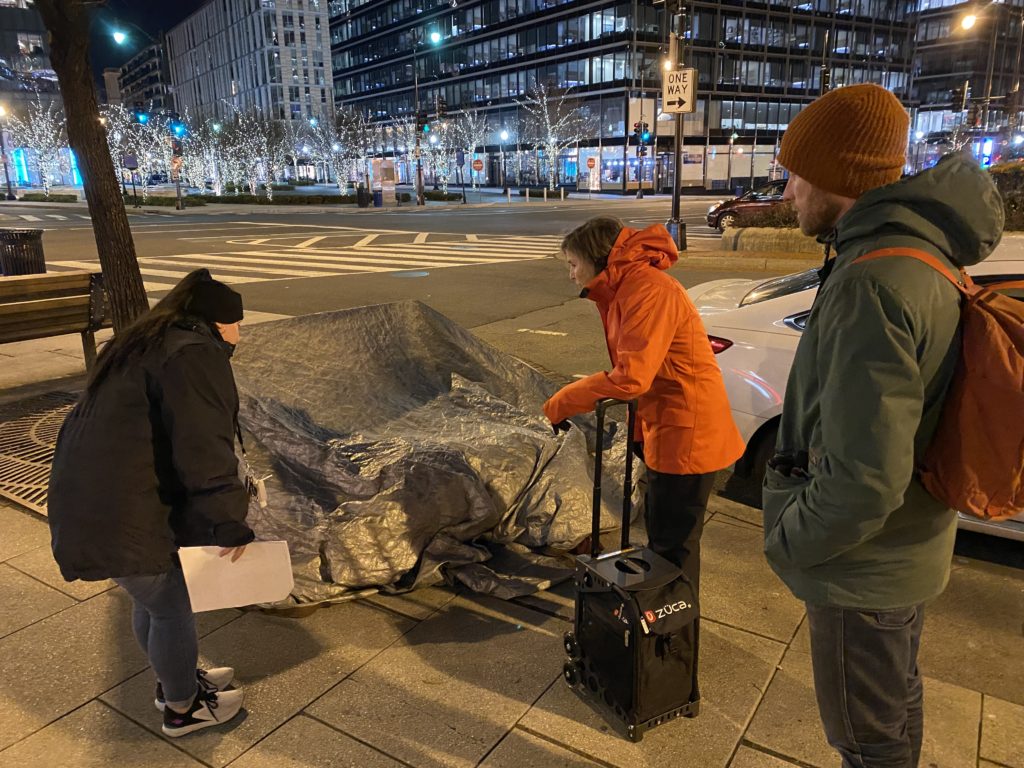 Three street outreach workers stand around a gray tarp concealing a homeless man. A Latina woman wear a black coat and leans down to talk to the man. A white woman in a puffy orange coat carries a black rolling bag of medical supplies. A tall white man in a green coat and wearing an orange backpack looks on.