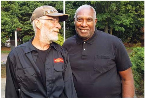 An older black man wearing a black shirt smiles. An older white man wearing a black shirt with a red tag and a green baseball cap with pins on it smiles with his head turned towards the other man.