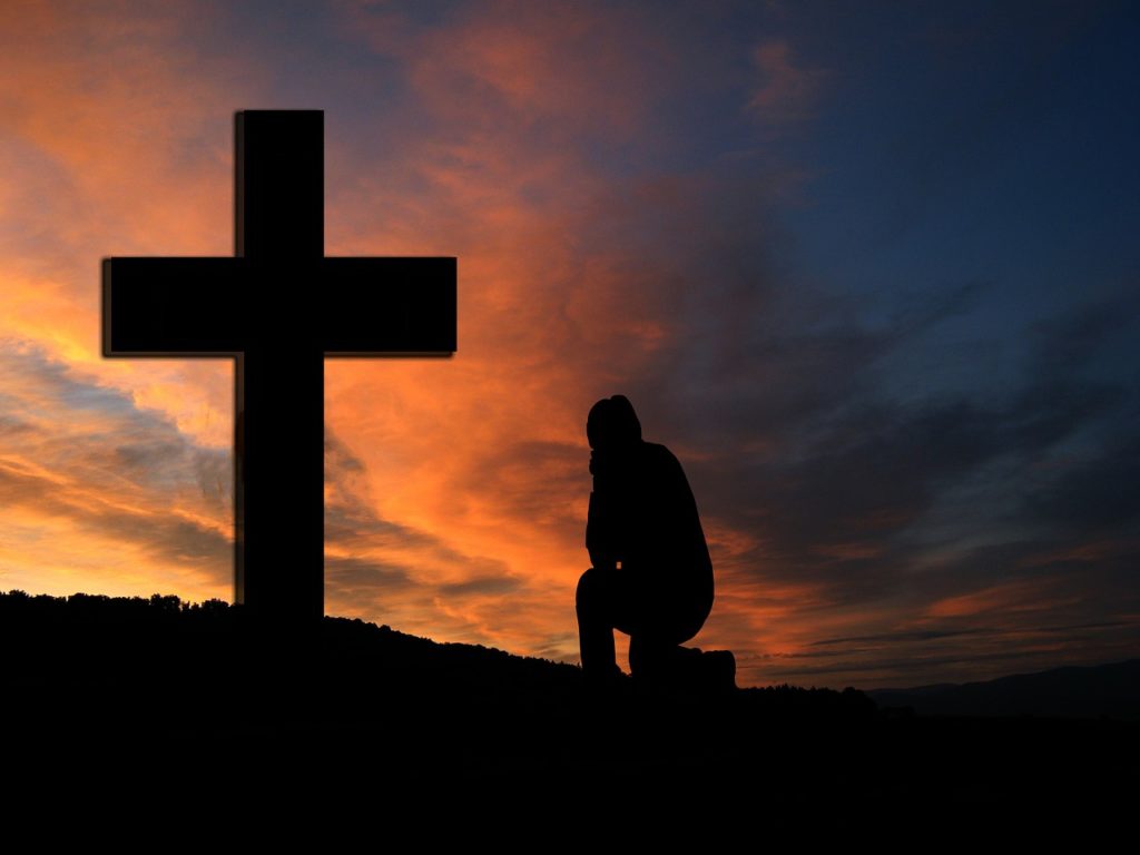 The sun sets in the sky, and a man on a hill kneels in front of a cross.