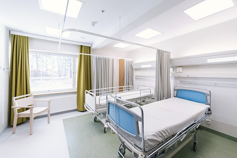Two hospital beds in a hospital room. An open window lets sun shine through. A chair sits in the corner.