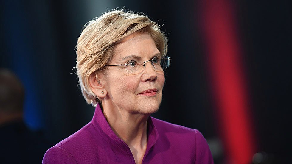Wearing a purple suit and glasses, Elizabeth Warren looks at the camera.