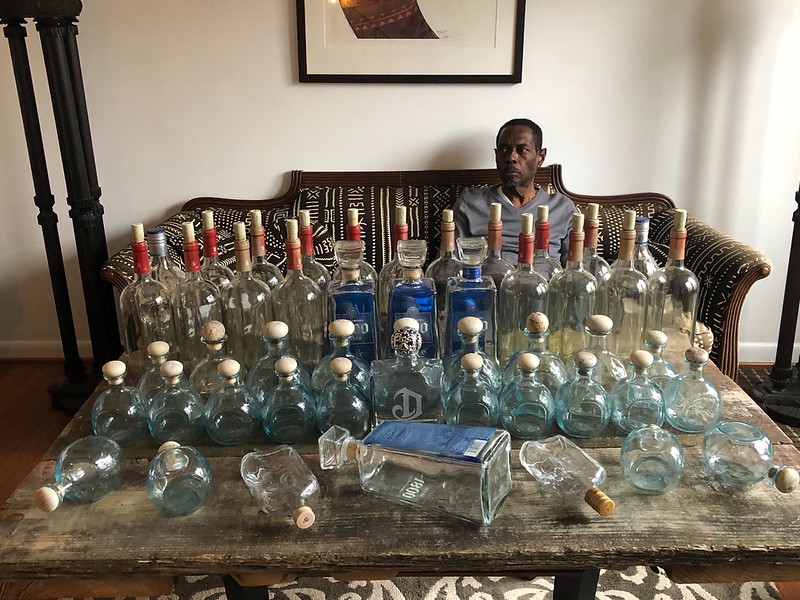 A photo of a man sitting in his living room in front of his artwork which consists of glass bottles of different sizes.