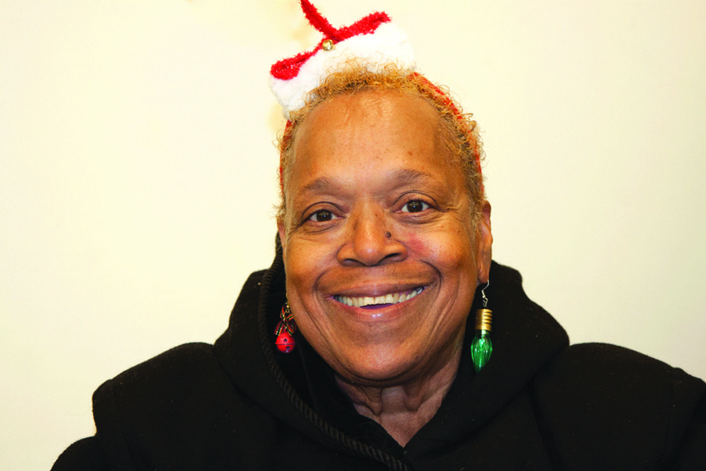 Photo of a black woman smiling. She wears a black sweatshirt, a small Santa hat and earrings shaped like Christmas lights.