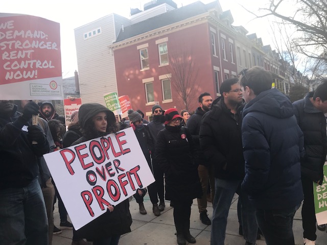 A woman in a hooded coat holds a sign that says "People over Profit" A crowd of people stand behind her. Everyone is in front of an apartment building