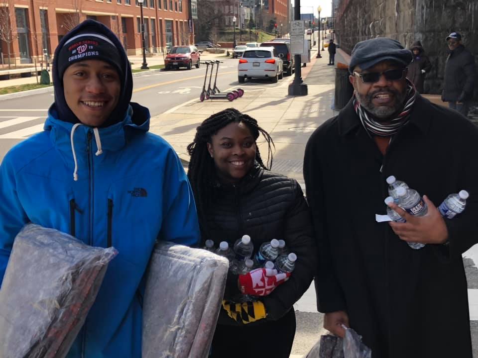 A tall black teenager holds blankets wrapped in plastic and wears a blue coat. A shorter black girl stands next to him holding water bottles. A black man in a black jacket stands next to her also holding water bottles. All are smiling and are standing on a street corner.