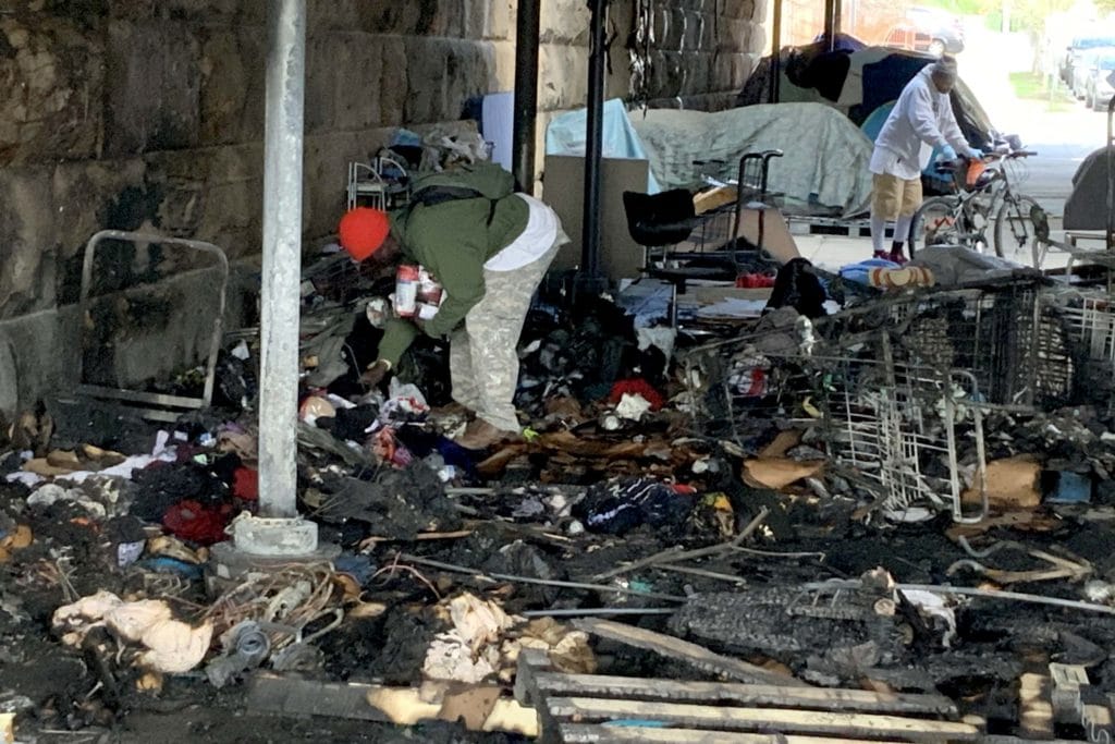 Photo of a man in a red hat bending over to examine the burned and now soaking wet remnants of his belongings.