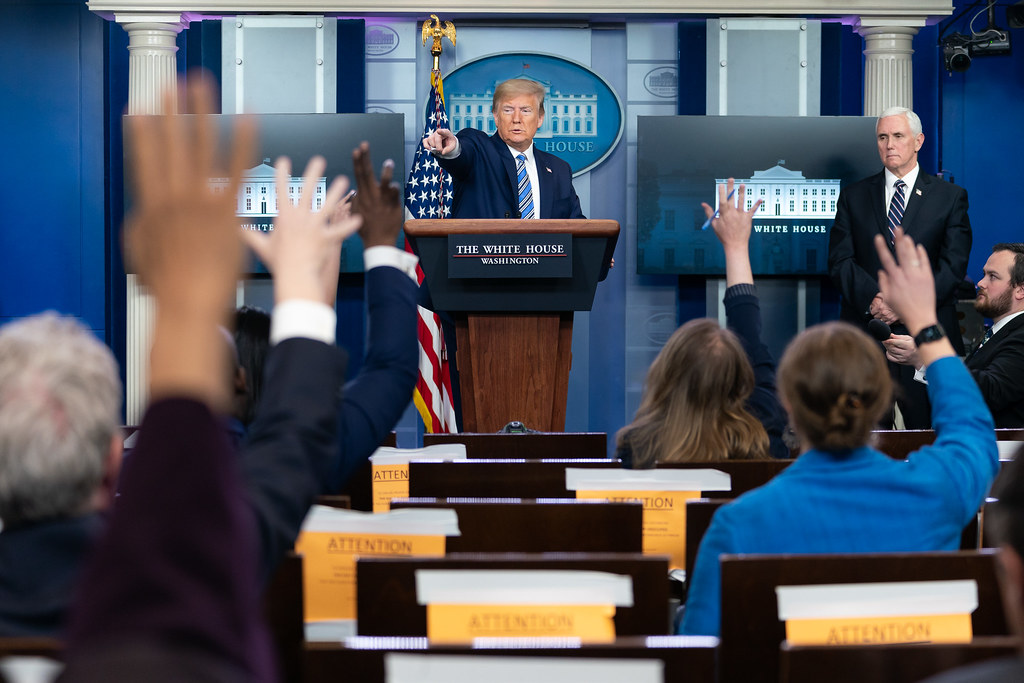 Photo of Trump conducting a White House press briefing. Trump is pointing towards a group of reporters with their arms raised.