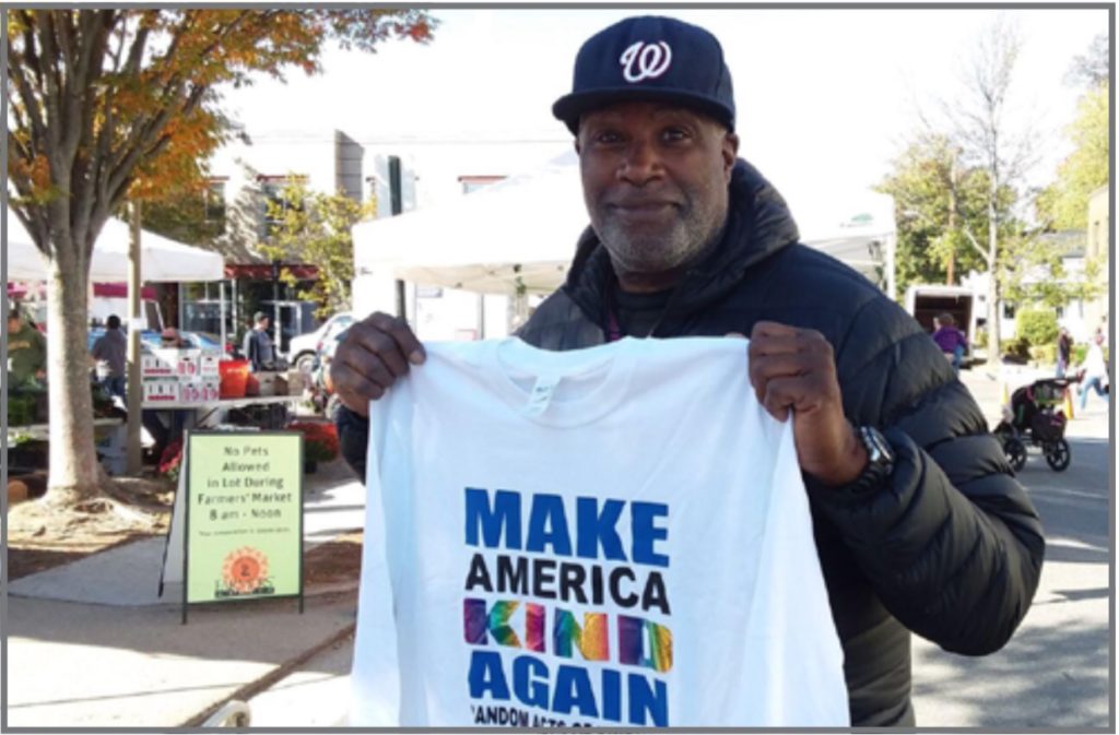 Photo of black man in in a black puffer jacket and a black Washington Nationals baseball cap. Man is holding a white t-shirt with the words make America kind again