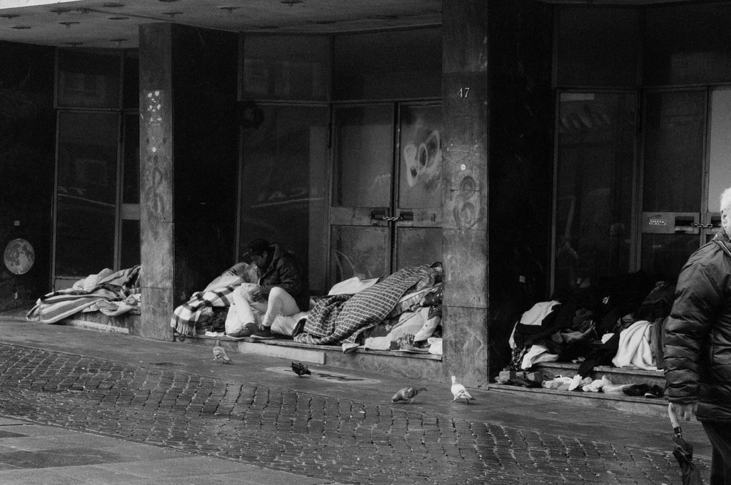 A black and white photo of a homeless encampment. Piles of blankets with people under them lie on the side of the street under a bridge.