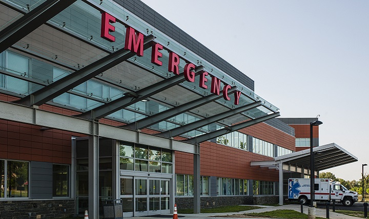 An ambulance sits outside a hospital. The hospital is made of glass and red brick. Emergency is spelled out in red lit up letters