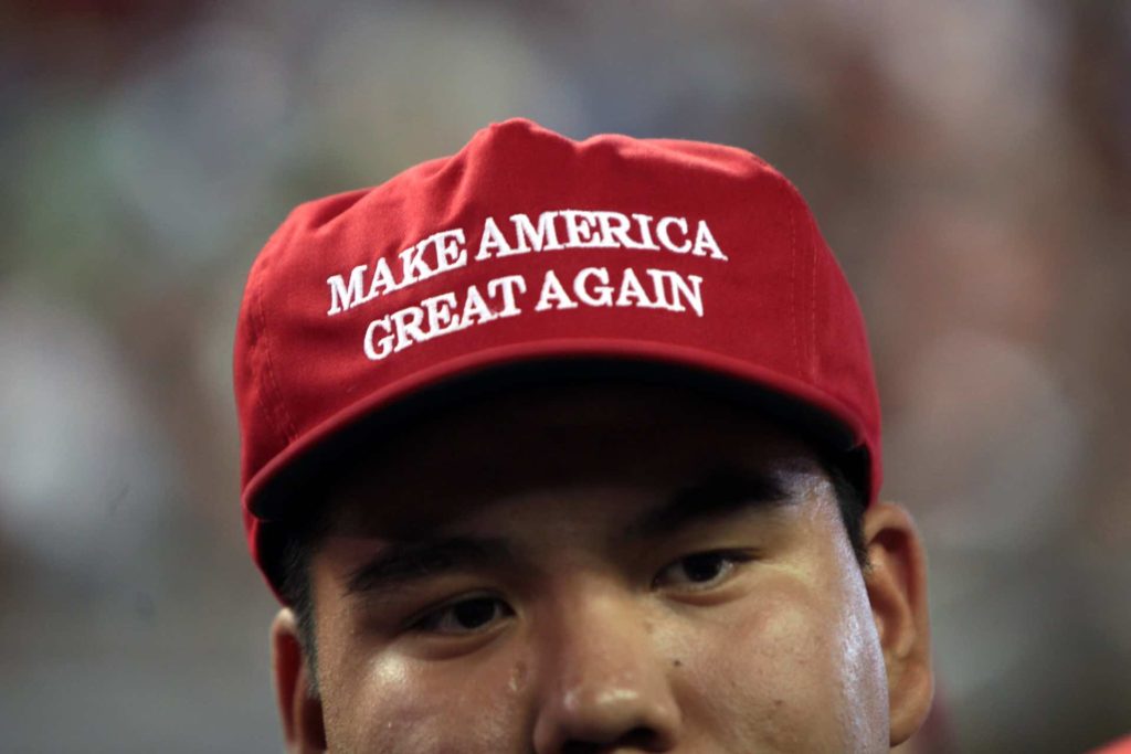 A close up photo of the top half of an Asian man's face. He wears a red hat with Make America Great in white text