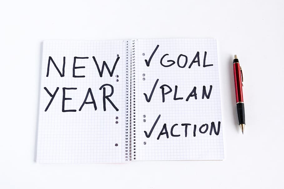 A photo of a lined-page notebook. The left side says New Year and the right side says Goal, Plan, Action with check marks next to each word. A red pen is on the right side of the notebook.