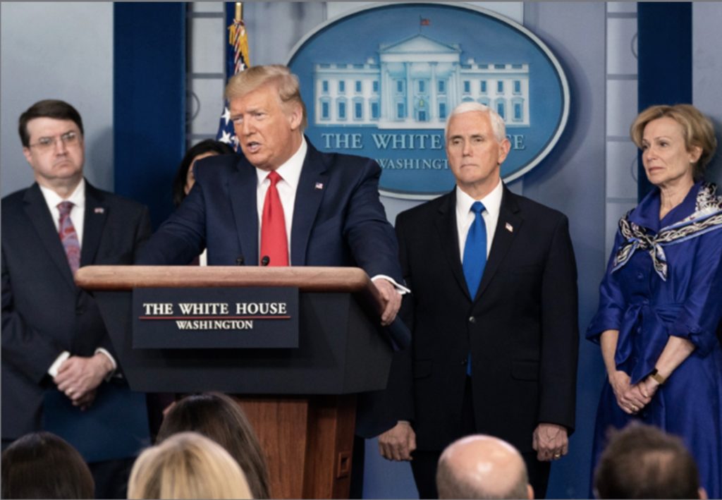 President Trump, a white man with blond hair stands behind a podium wearing a blue suit and a red tie.