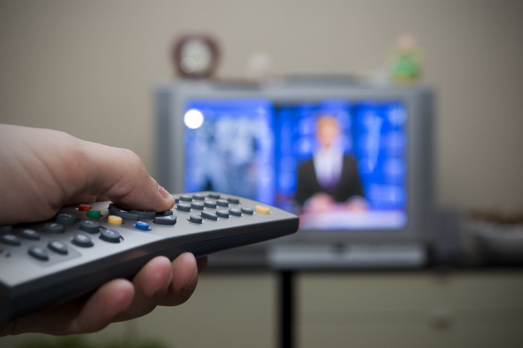 A hand holds a tv remote. A tv is blurry in the background depicting a tv anchor in front of a blue background