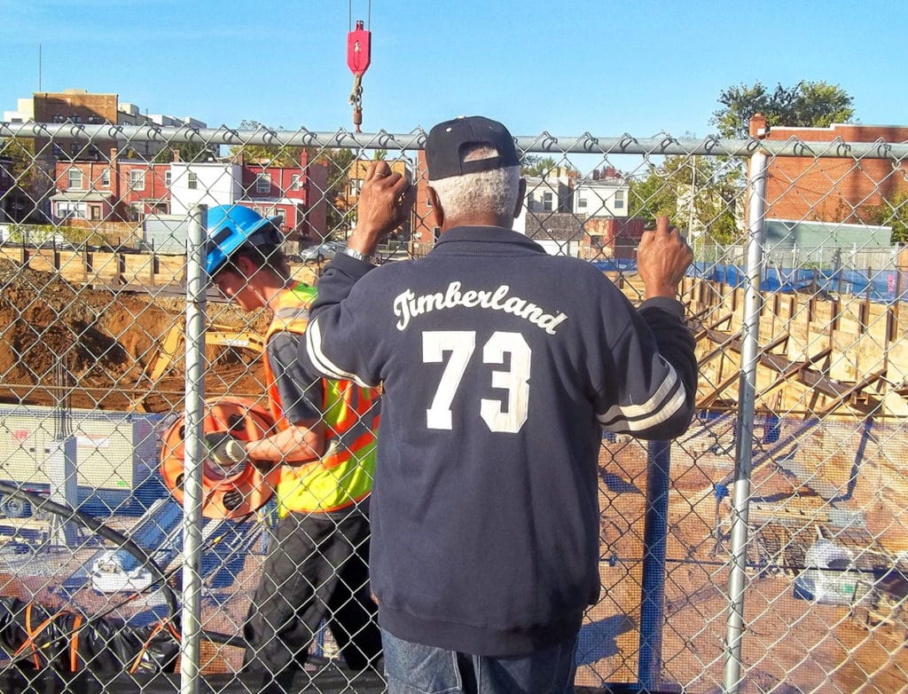 A black man in a navy jacket that has the number 73 and the word Timberland stitched onto it looks through a fence. His back is to the camera. A white young man holds construction cable in a construction site on the other side of the fence.