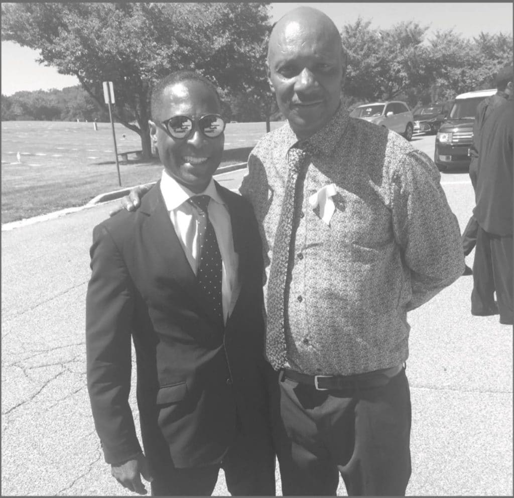 Two black men stand next to each other smiling at the camera. The man on the left wears a suit and tie. The man on the left wears a striped button up shirt and a tie