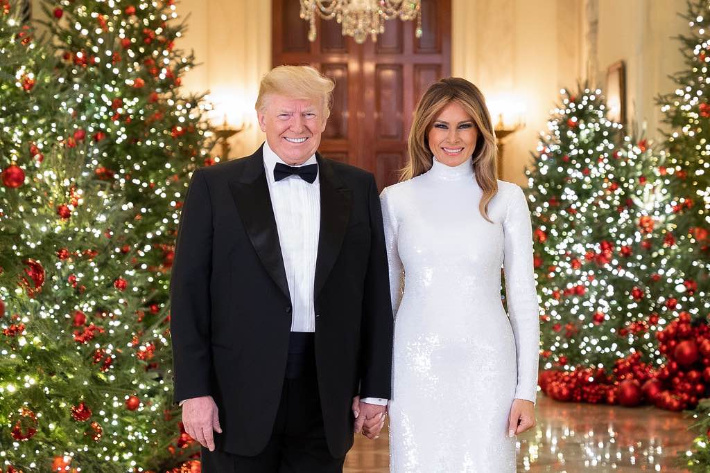 President Donald trump wears a tuxedo his wife to his wife wears a white tight skin dress. Christmas trees are in the background.