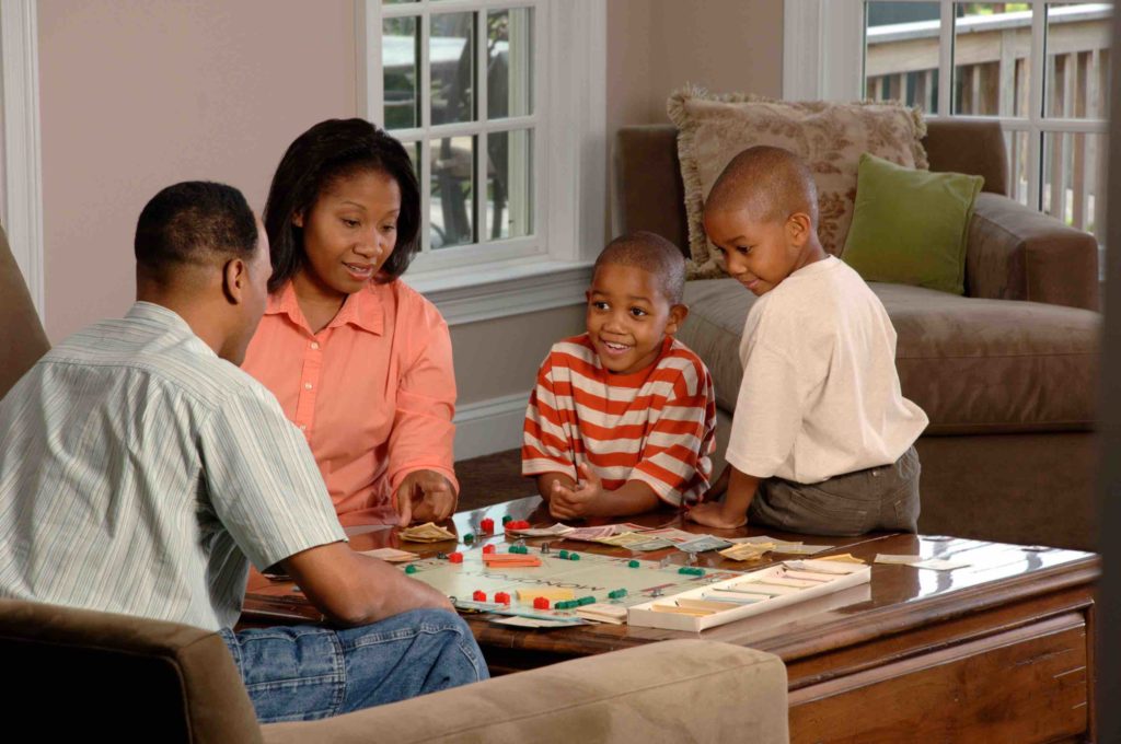 A black family playing a board game. The family is made up of a mom in an orange shirt and two young boys and a dad wearing a blue button up. One boy wears a striped red and white shirt and the other boy wears a white t-shirt.