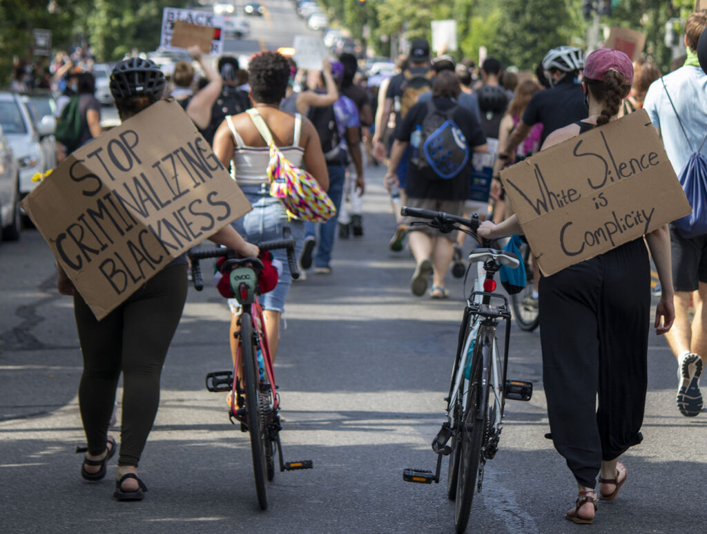 Photo of protesters walking their bikes carrying signs that say "stop criminalizing blackness" and 'white silence is complicity", with more protesters marching in front of them.