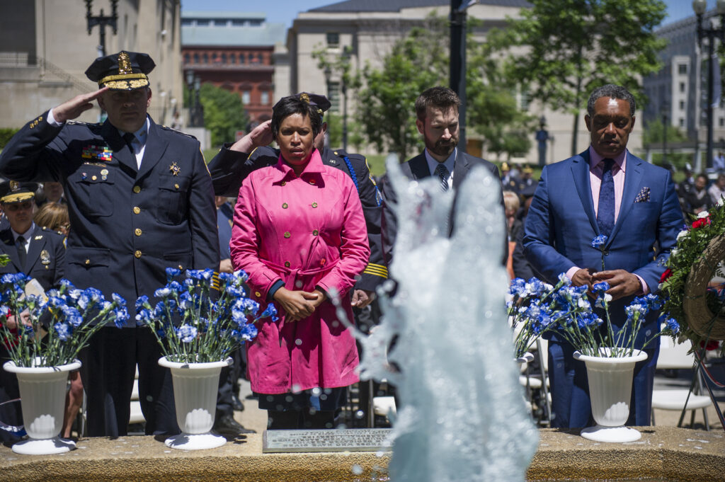 Mayor Bowser stands next to police officials behind flowers and a fountain.
